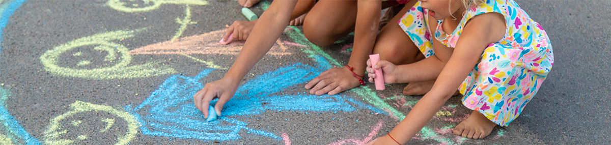 Das Bild zeigt zwei Kinder im Sommer, die mit bunter Kreide Figuren auf einen betonierten Boden malen.